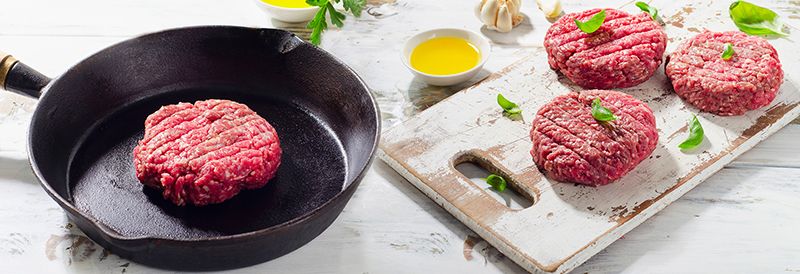 Drei Rohe Burgerpatties angerichtet auf einem Brett. Ein Patty ist in einer Pfanne