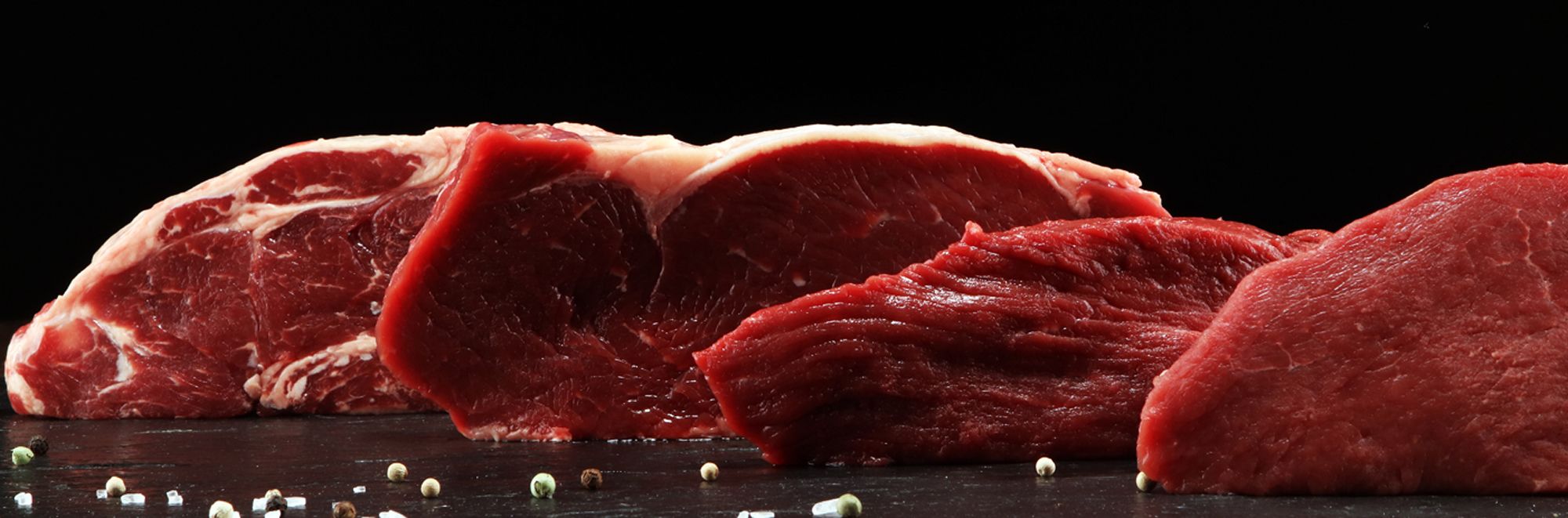Rohe Rindfleischstücke vor dunklem Hintergrund. Ein paar Pfefferkörner liegen auf dem Tisch.