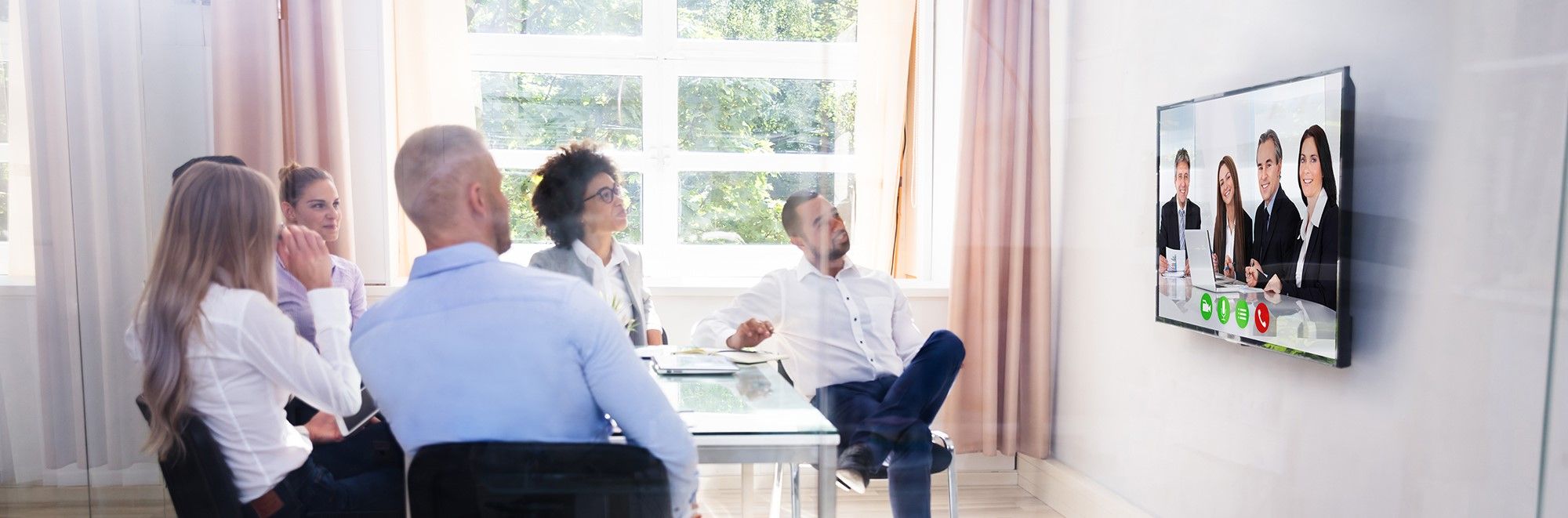 Mehrere Personen sitzen in einem Konferenzraum. Auf einem Bildschirm sind weitere Personen zu erkennen